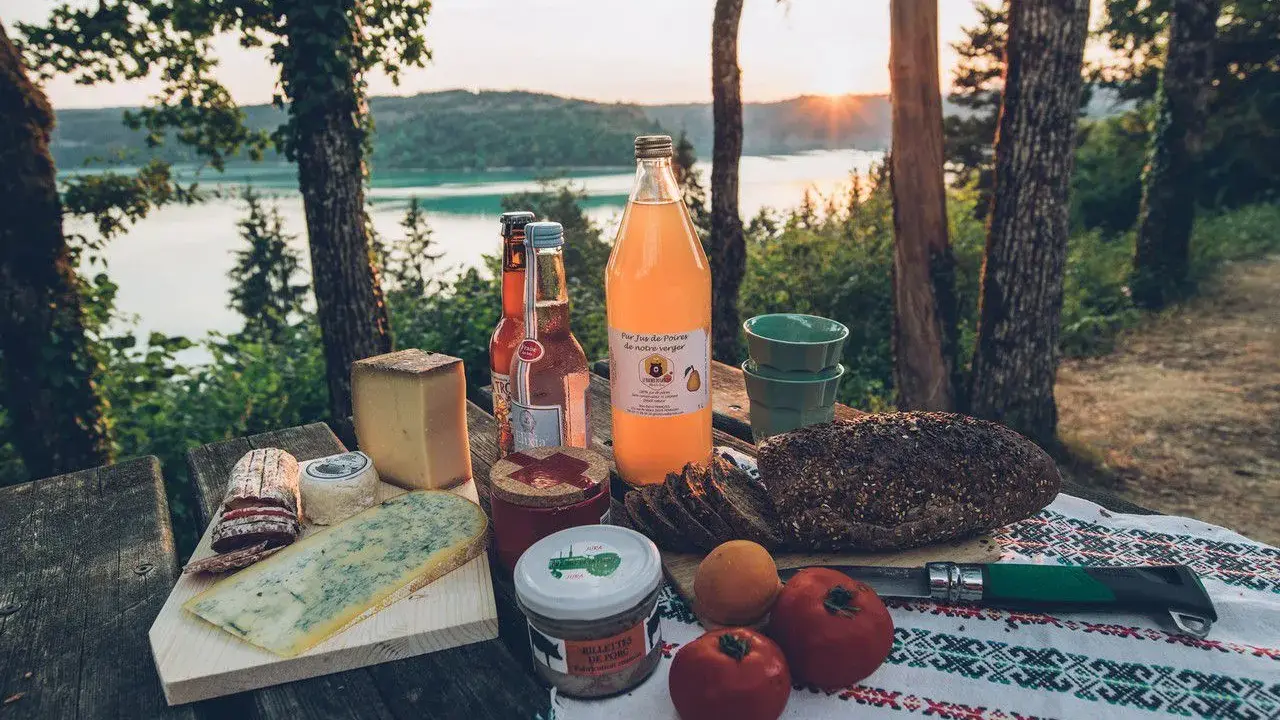 Pique-nique avec fromages, pain de campagne, jus de poire et terrine, un aperçu des spécialités culinaires Jura au bord d'un lac.