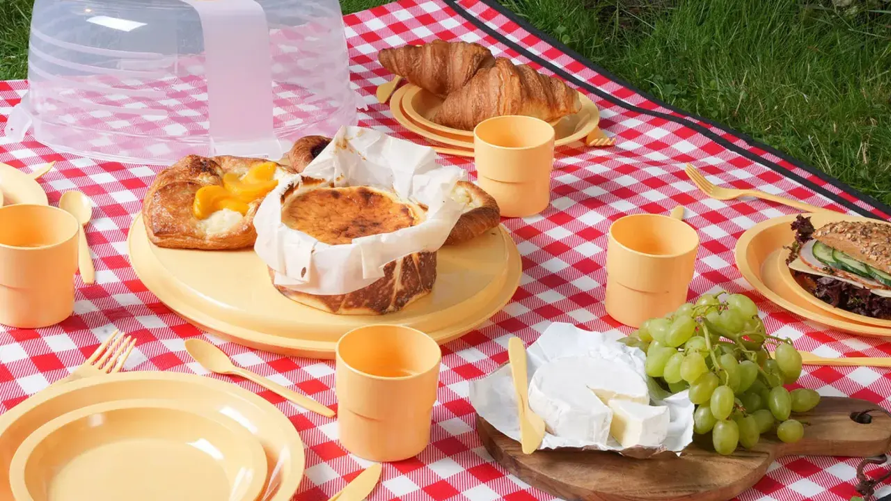 Co zabrać na piknik? Na czerwono-białej kratce leżą ciasta, croissanty, ser, winogrona i kanapka. Są też żółte talerze, kubeczki i sztućce.
