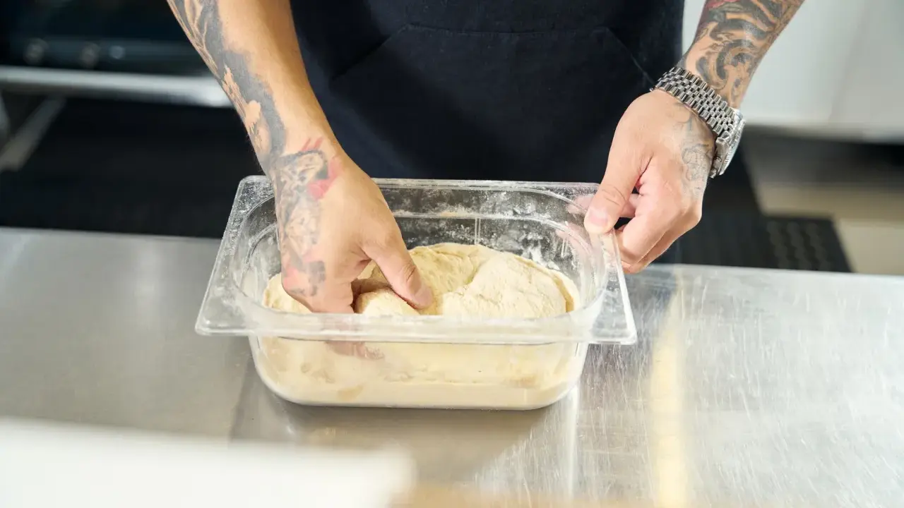 Ręce z tatuażami zagniatają ciasto w plastikowym pojemniku. To pierwszy krok, by dowiedzieć się, jak mrozić ciasto.