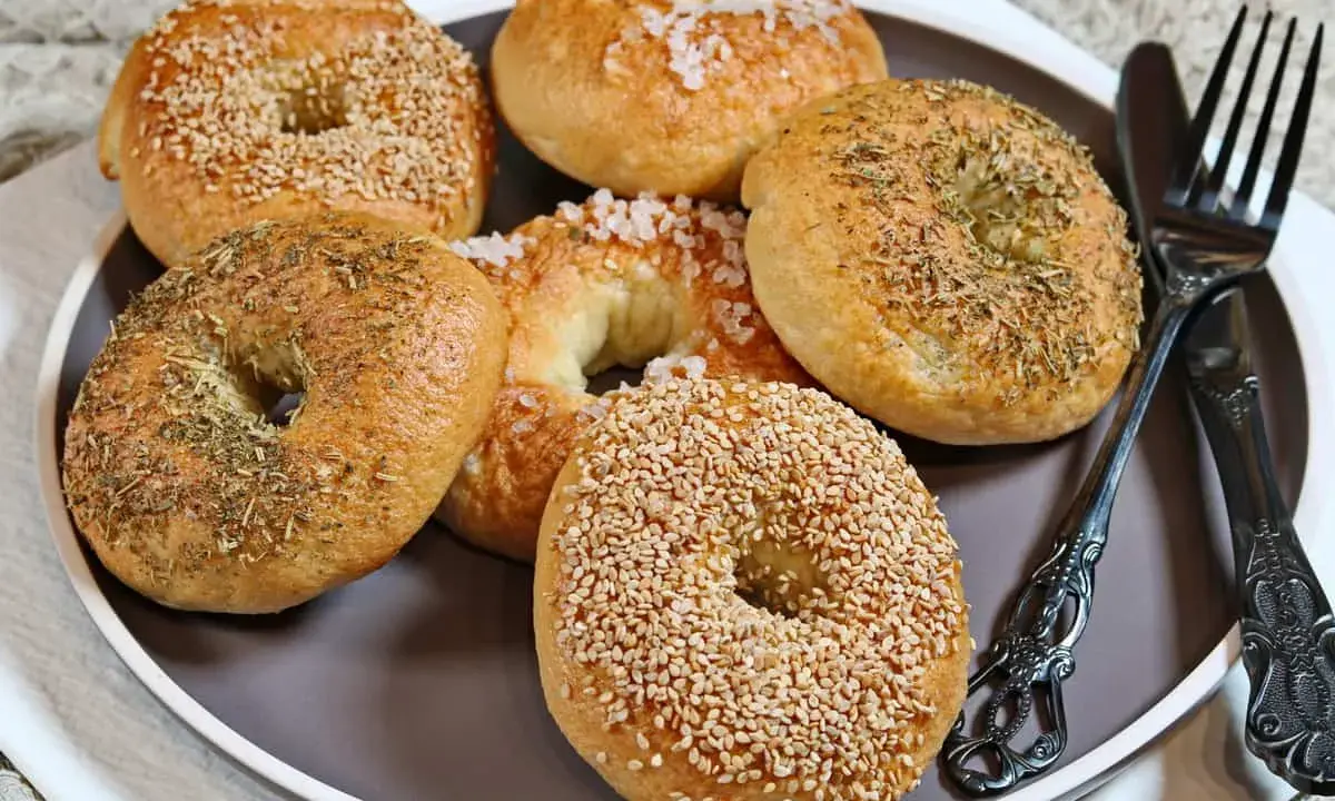 Sechs selbstgemachte Bagels auf einem Teller: zwei mit Sesam, zwei mit Kräutern und zwei mit grobem Salz bestreut. Ein Messer und eine Gabel liegen daneben.