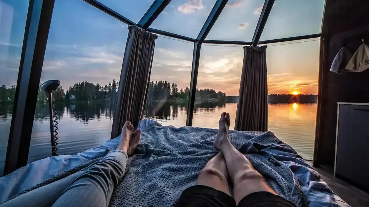 Två personer vilar i en glaskupa med utsikt över en sjö i Norrland vid solnedgången. En perfekt glampingupplevelse.