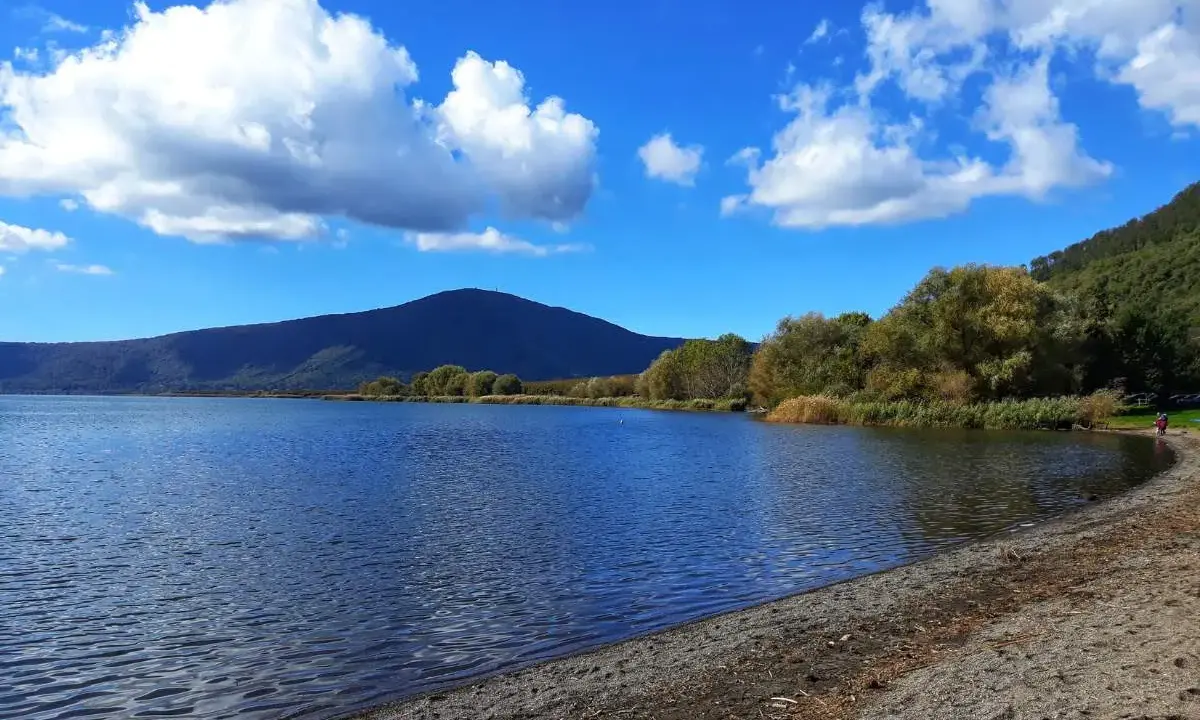 Spiaggia ghiaiosa e acque calme del lago di Vico, con montagne verdi e cielo azzurro punteggiato di nuvole bianche.