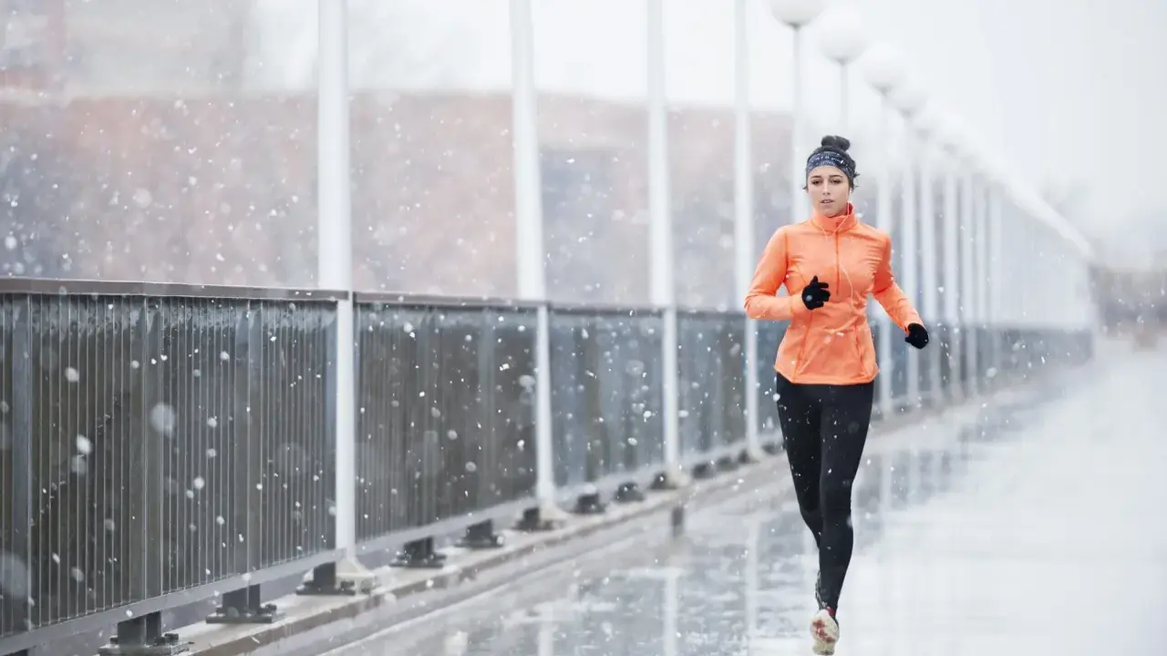 Donna corre sotto la pioggia su un ponte, con fiocchi di neve che cadono. Indossa abbigliamento sportivo arancione e nero.