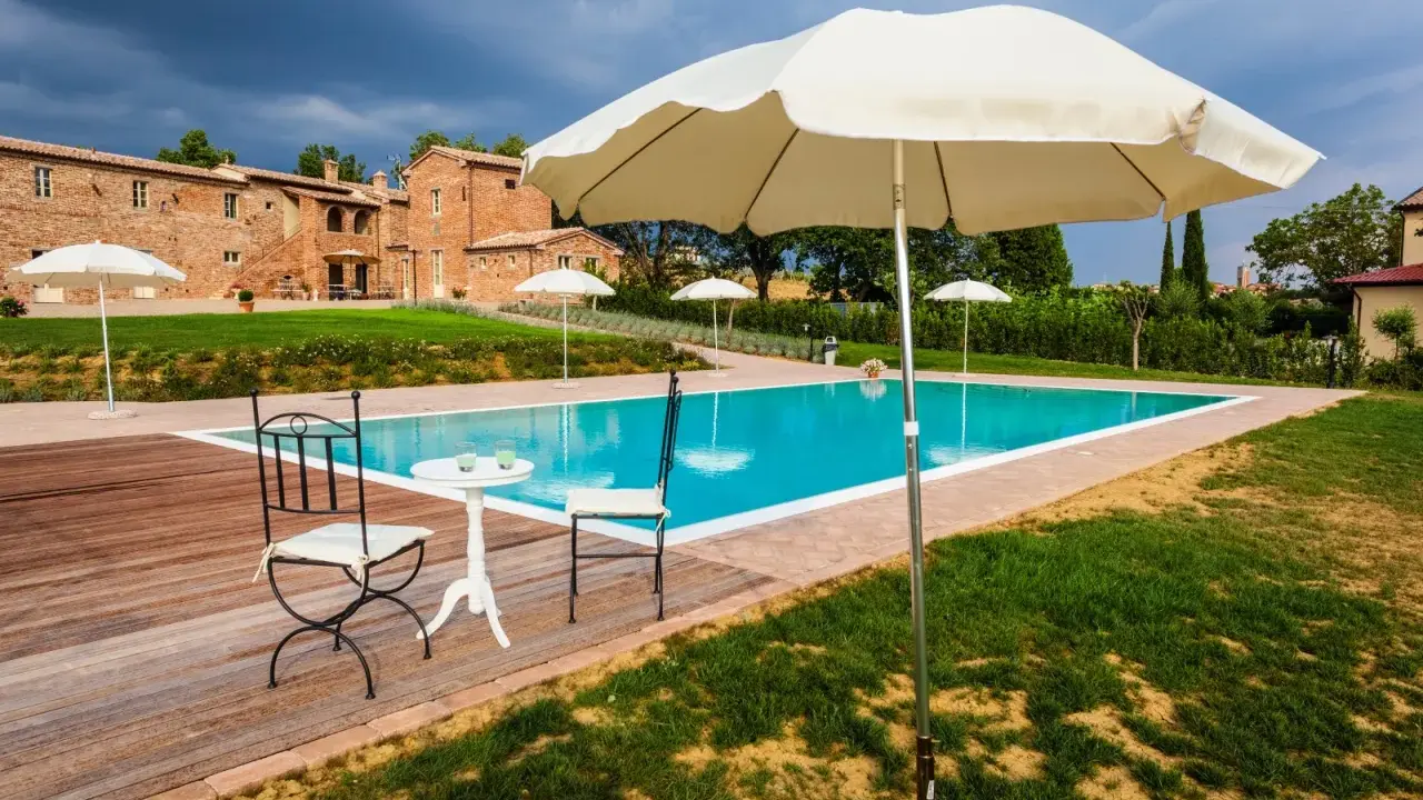 Piscina con vista su un casale in mattoni, perfetta per le tue vacanze in Toscana. Tavolino con sedie e ombrelloni pronti per rilassarsi.