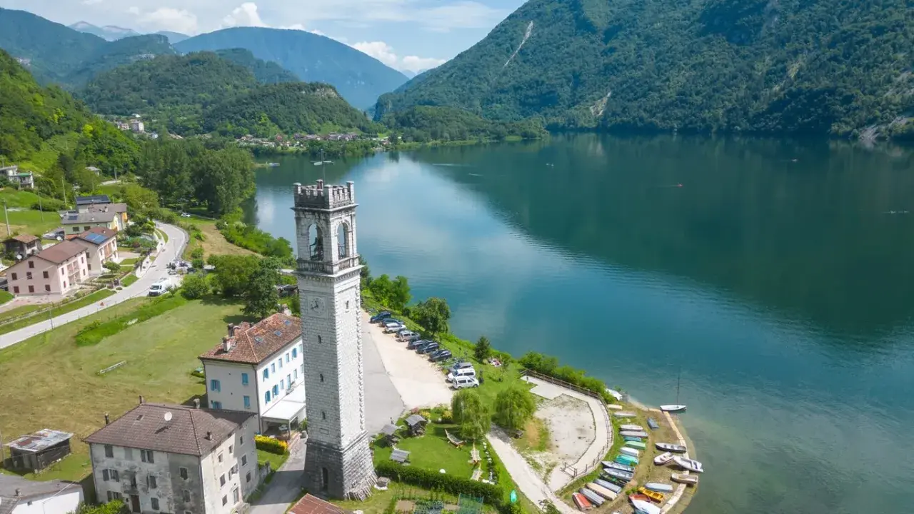 Torre campanaria affacciata sul lago di Corlo, con montagne verdi sullo sfondo e un villaggio pittoresco.