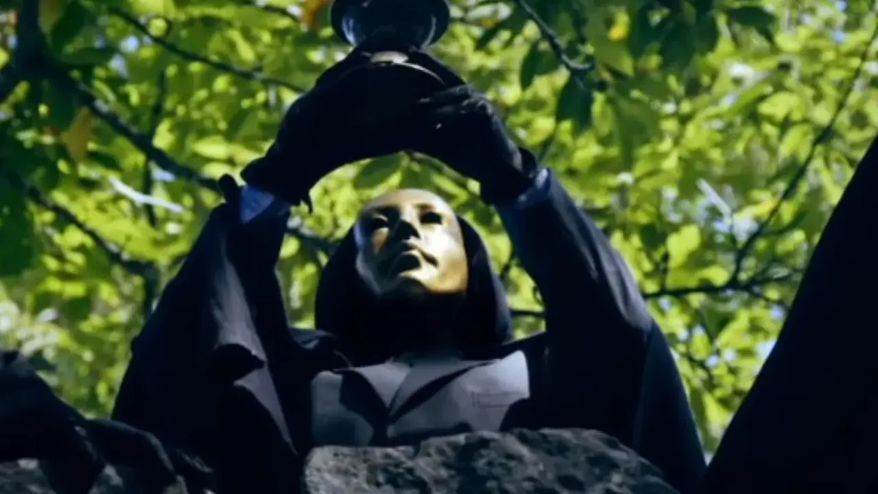 En figur med gyllene mask och svart kappa håller upp en bägare mot lövverket. Kanske är det "svarta solen geografens testamente" som avslöjas.