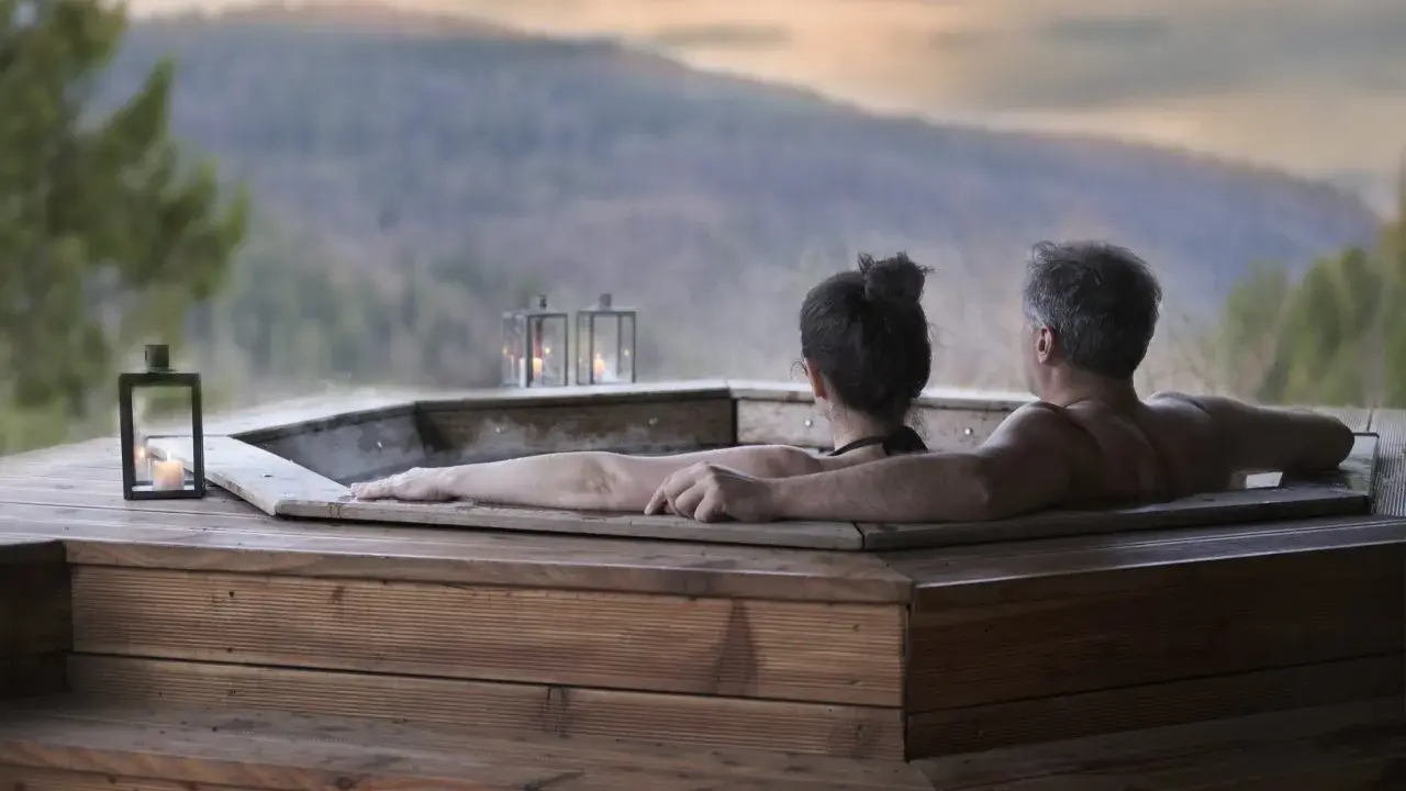 Coppia abbracciata in una vasca idromassaggio di legno, con lanterne accese e vista sulle montagne. Un momento di puro relax in Trentino.