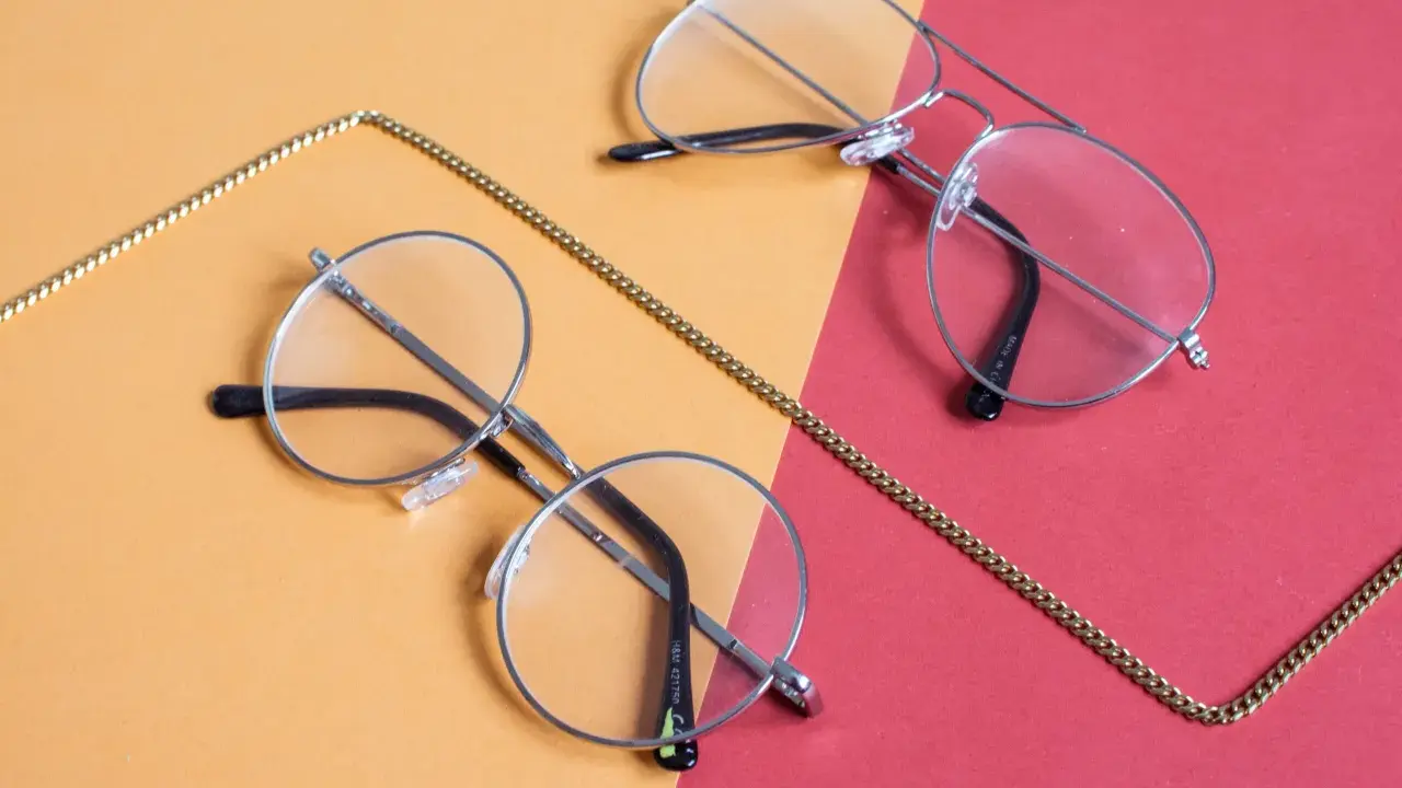 Dwa stylowe okulary, okrągłe i aviator, leżą na pomarańczowo-czerwonym tle. Złoty łańcuszek oplata je, sugerując, że to okulary korekcyjne, być może na astygmatyzm.