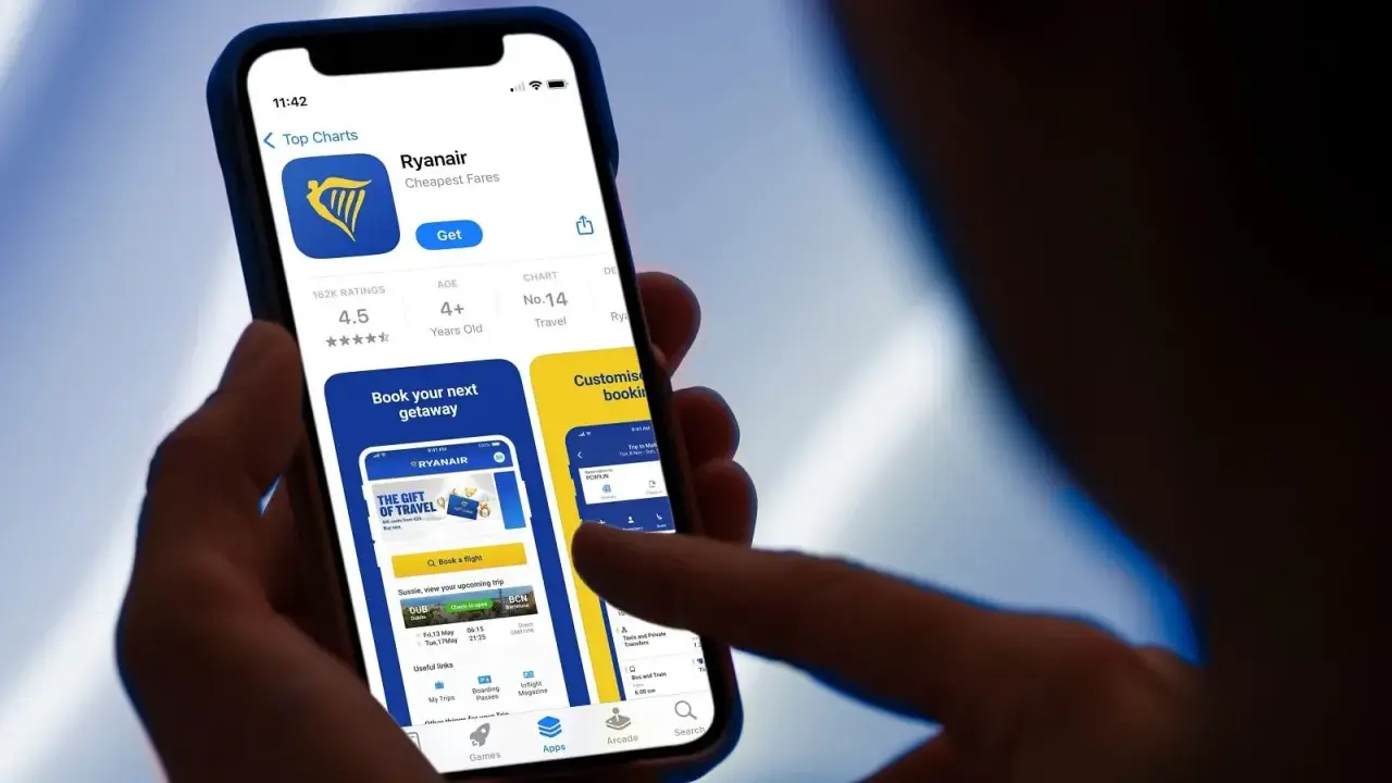 Dłoń trzyma smartfon z otwartą aplikacją Ryanair, widoczna jest strona główna z opcją rezerwacji lotów.