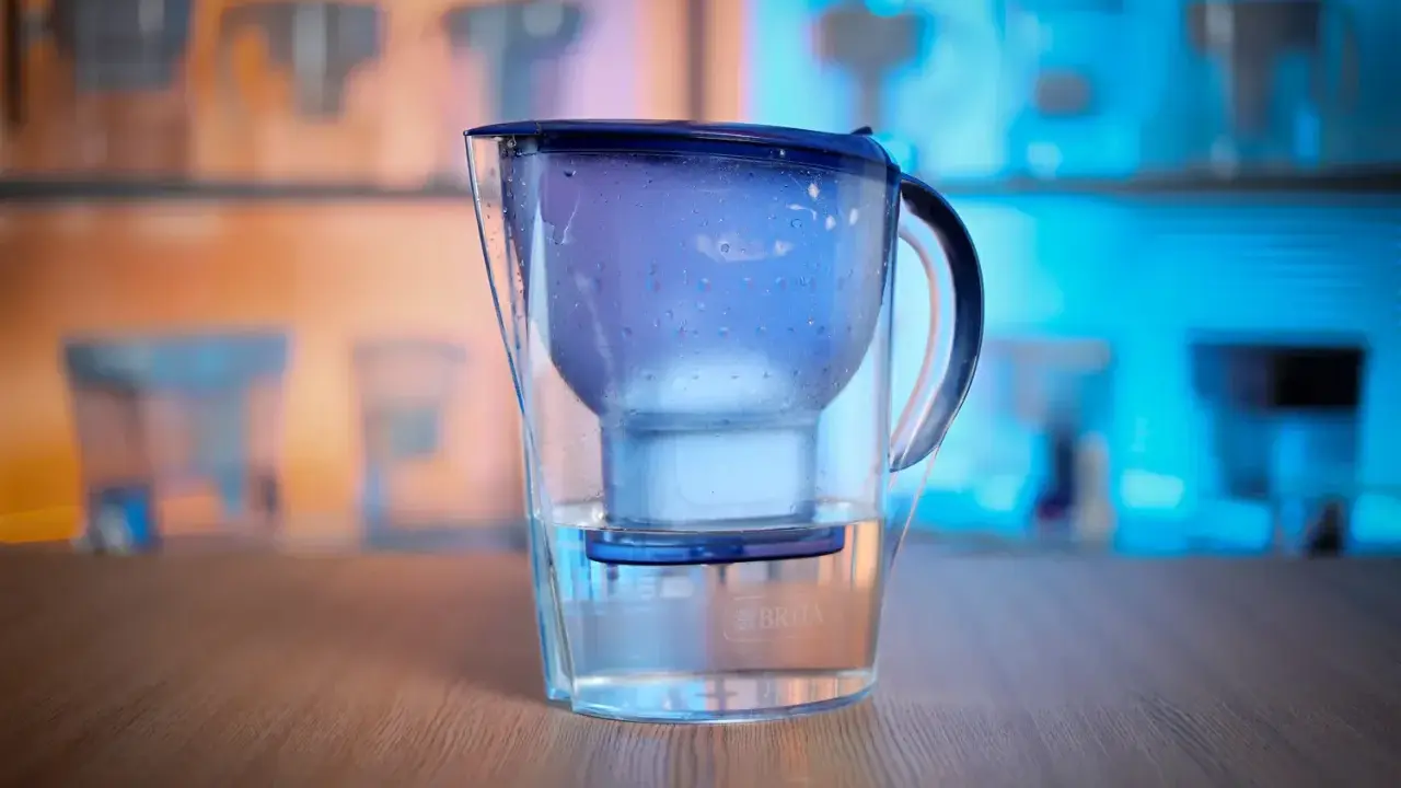 En vattenrenare för dricksvatten, en Brita-kanna, står på ett träbord. Bakgrunden visar fler liknande kannor i blått och orange ljus.