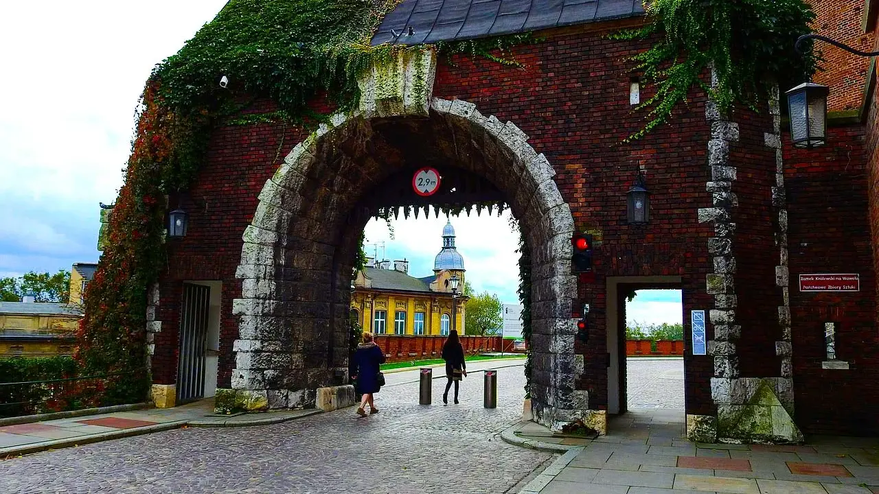 Brama na Wawel, gdzie można sprawdzić wawel bilety cennik. Kamienna konstrukcja porośnięta bluszczem, z widokiem na dziedziniec.