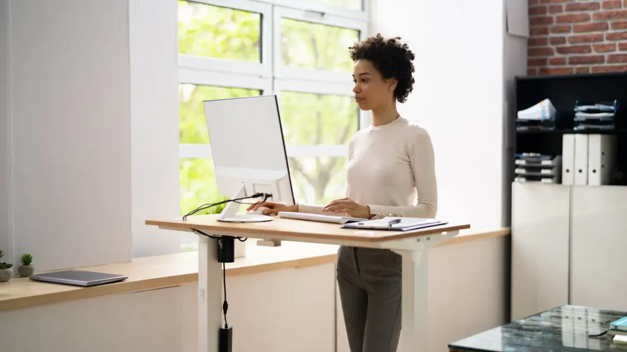 Une femme travaille sur un ordinateur à un bureau réglable en hauteur. La position debout prolongée présente des risques pour la santé, il est donc important de varier les postures.