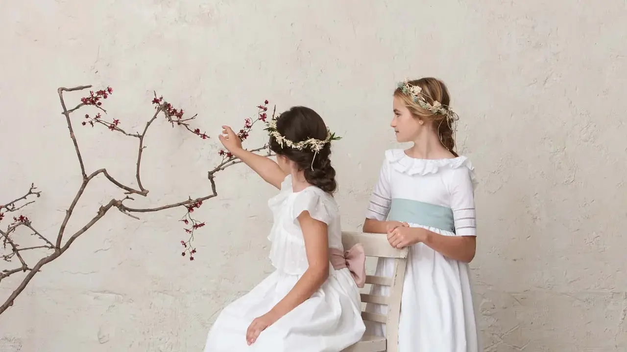 Dos niñas con vestidos blancos y coronas de flores, una sentada tocando una rama, la otra de pie. Peinados de trenza para niña de comunión.