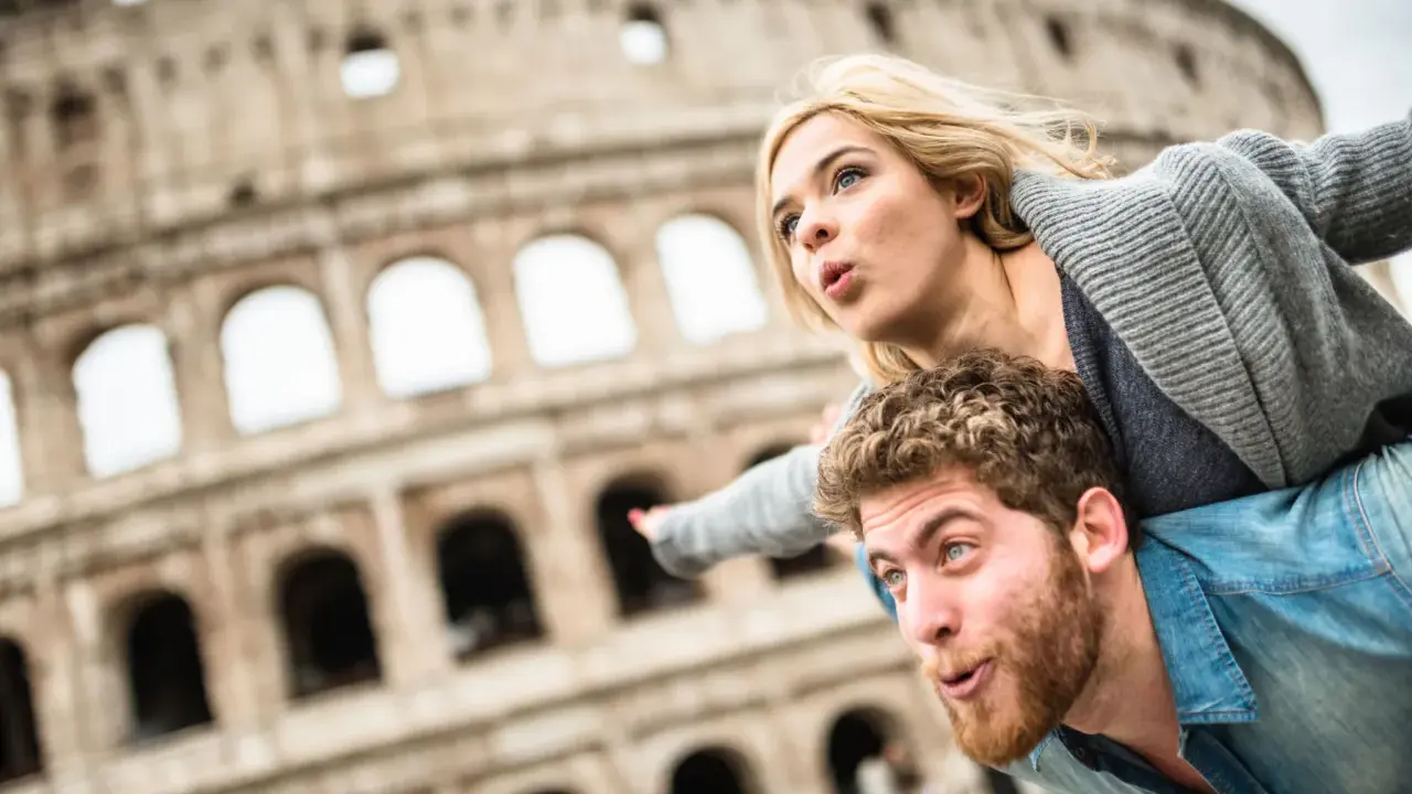 Coppia in una vacanza romantica a Roma, con il Colosseo sullo sfondo. Lui porta lei sulle spalle, entrambi con espressioni sorprese.