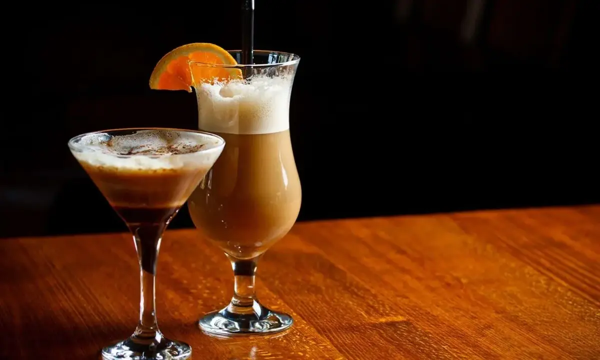 Dwa drinki z likierem kawowym i mlekiem: jeden w kieliszku martini, drugi w wysokiej szklance z pomarańczą.
