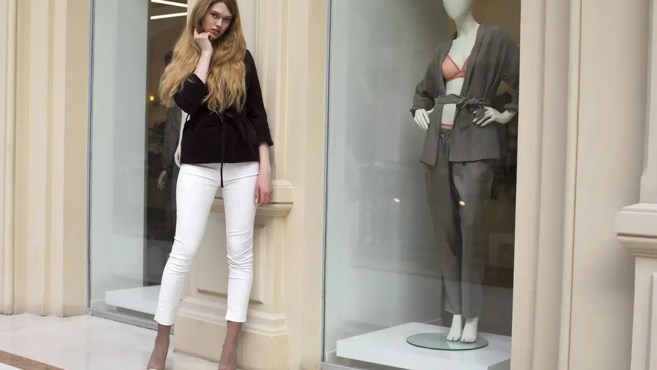 Modelka w białych spodniach i bordowej marynarce obok manekina w eleganckim stroju. Skandynawskie marki odzieżowe prezentują najnowsze trendy.