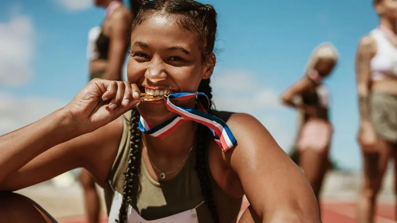 Läuferin beißt stolz in ihre Goldmedaille nach einem 10k Lauf. Andere Athletinnen im Hintergrund.