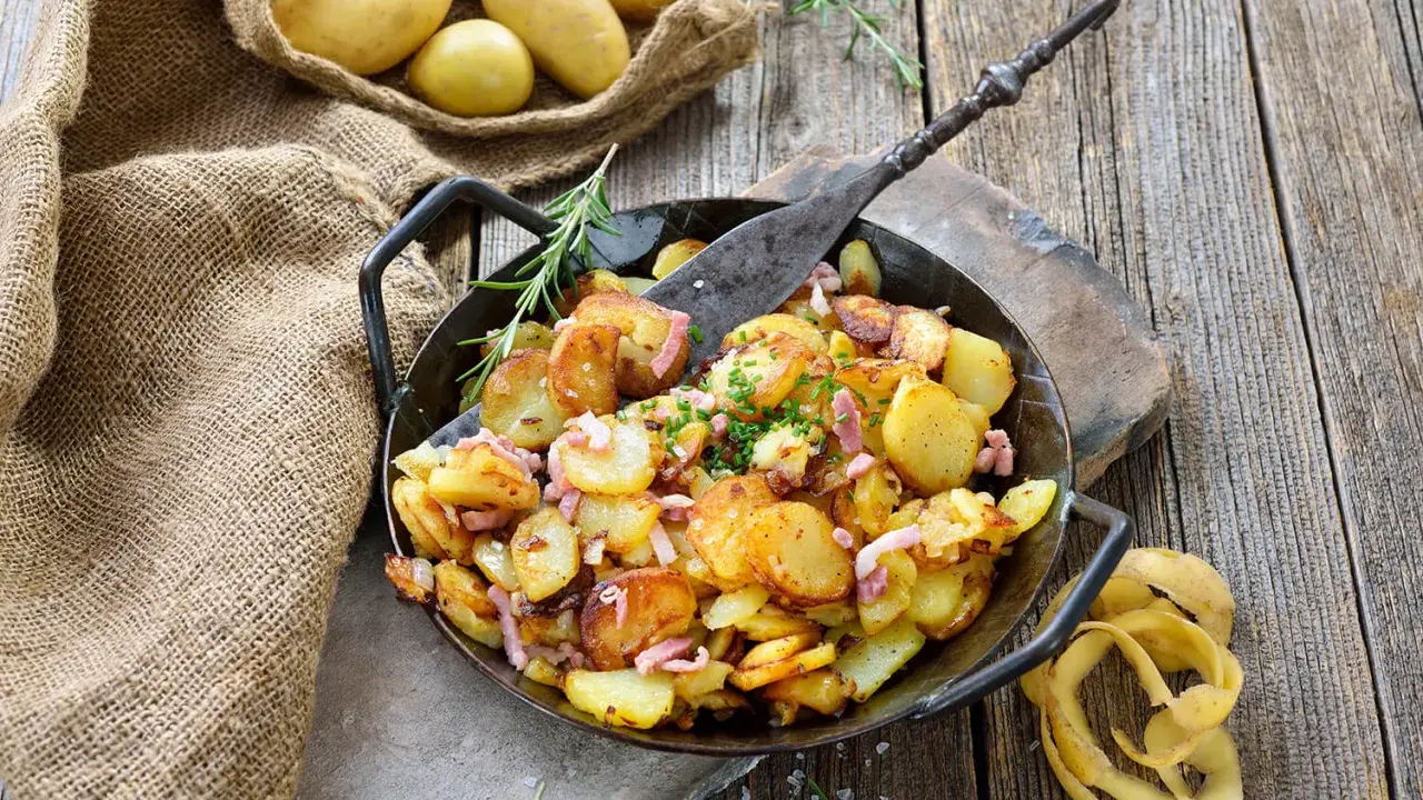Bratkartoffeln, die vorher gekocht wurden, in einer Pfanne mit Speck und Kräutern garniert. Daneben liegen rohe Kartoffeln in einem Jutesack.