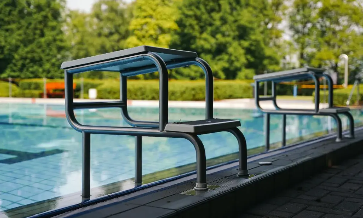 Zwei Sprungbretter am Beckenrand eines Freibades mit Bäumen im Hintergrund.