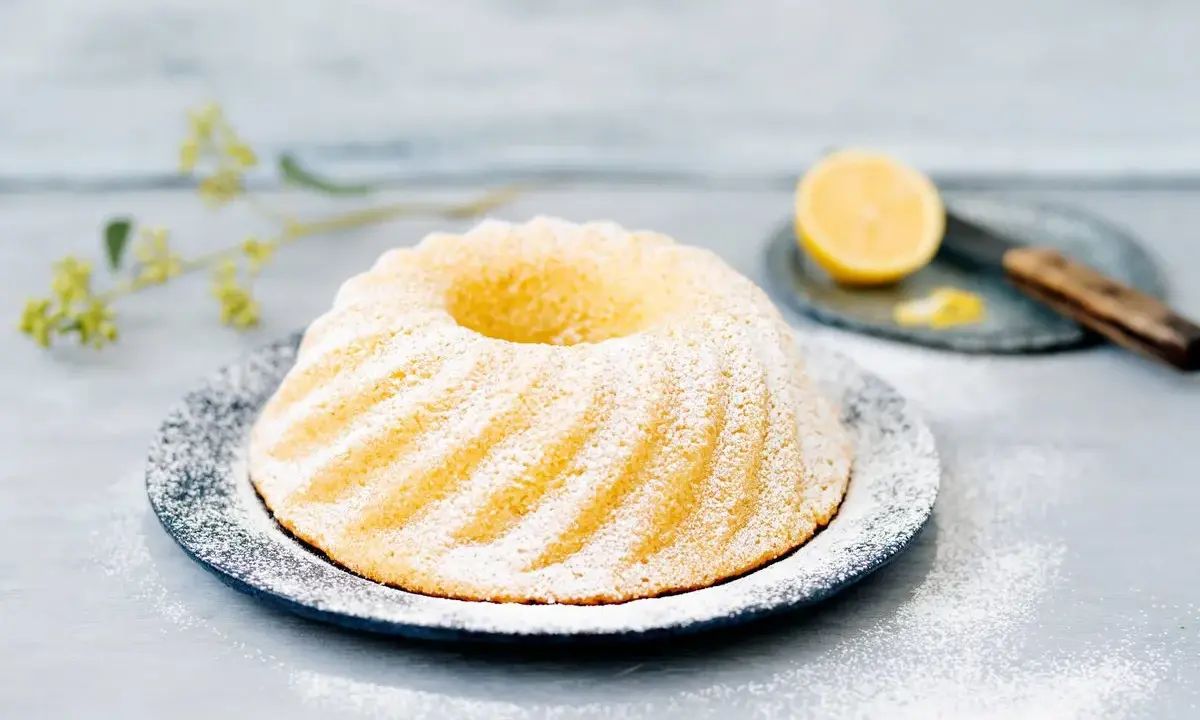 En saftig sockerkaka citron, pudrad med florsocker, serveras på ett fat. En halv citron och en kniv syns i bakgrunden.