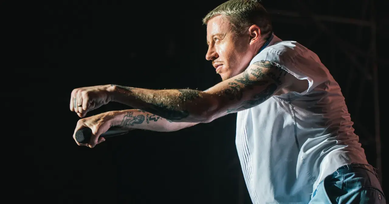 Macklemore na koncercie w Polsce, z mikrofonem w dłoni, w białej koszuli i z tatuażami na ramionach.