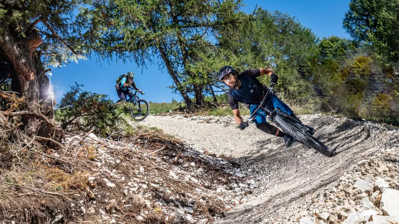 Due ciclisti affrontano un sentiero sterrato in discesa, immersi nella natura. La **tonezza del Cimone** si percepisce nell'aria fresca e nel paesaggio montano.