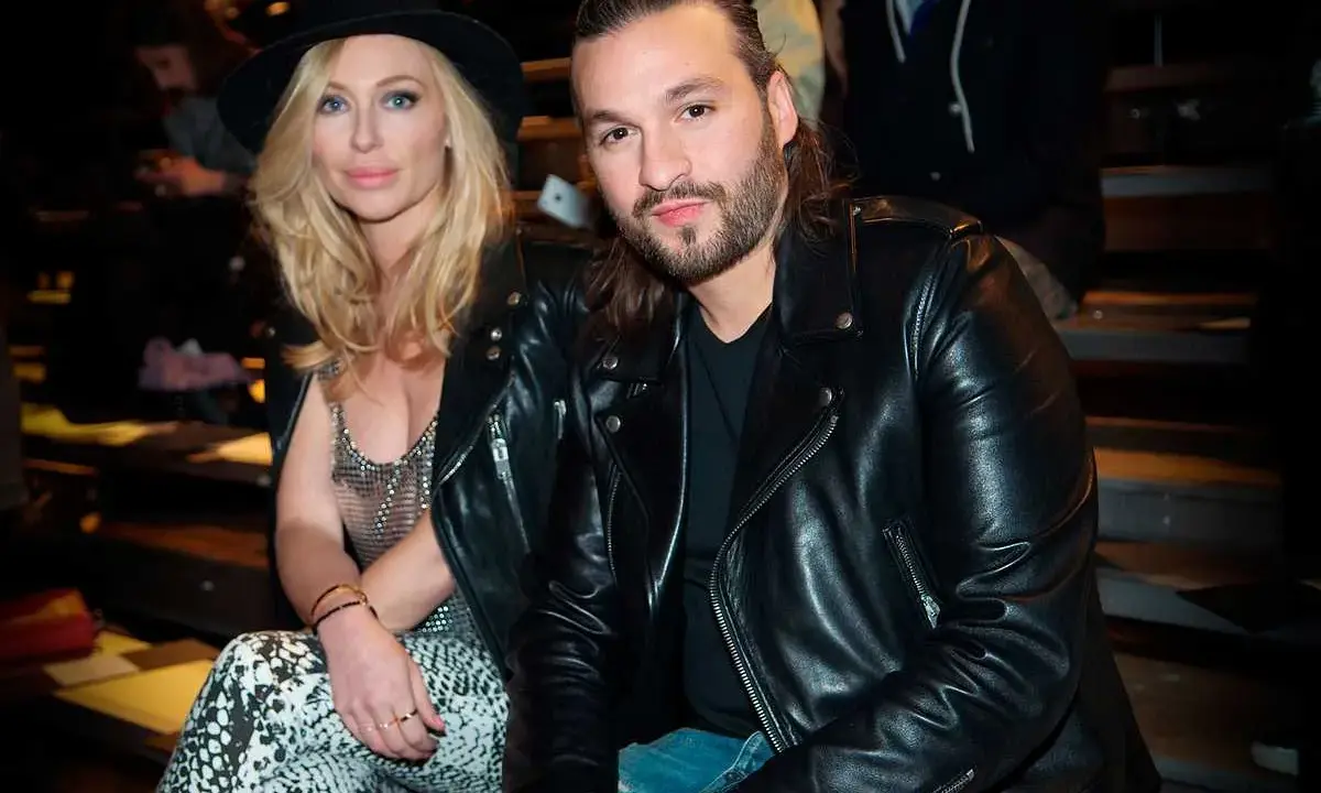 Steve Angello fru, Isabella, med sin blonda frisyr och hatt, sitter bredvid honom. Båda bär svarta läderjackor.