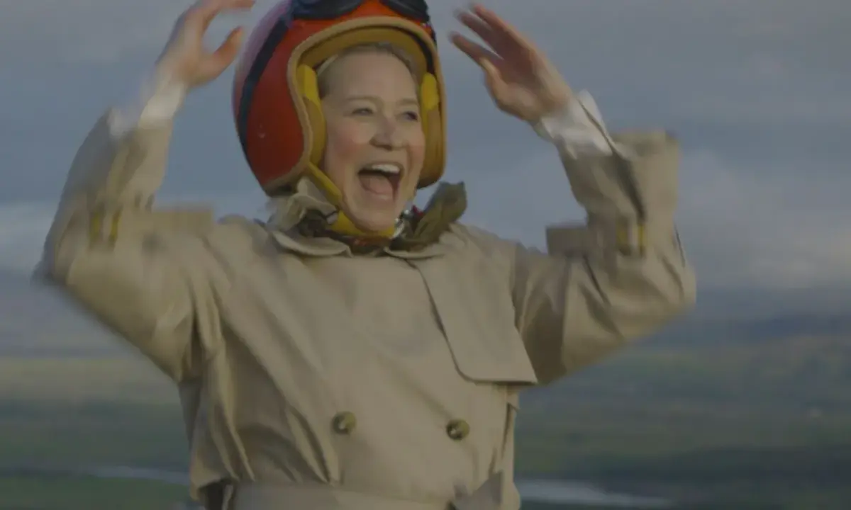 Glad Trine Dyrholm i pilotmössa och trenchcoat vinkar glatt. En scen från filmer med Trine Dyrholm.