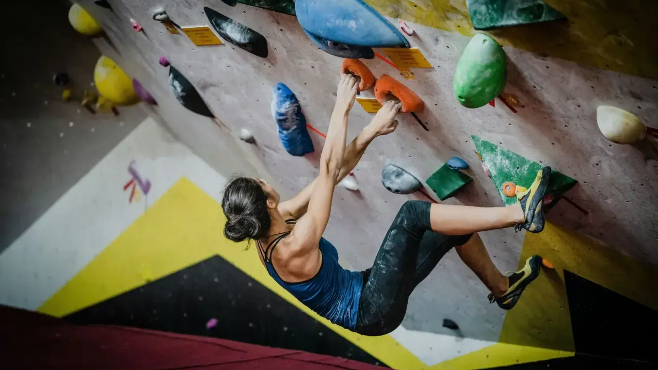 Kobieta wspina się po kolorowej ścianie, chwytając pomarańczowe uchwyty. To właśnie bouldering – sportowa wspinaczka bez liny.