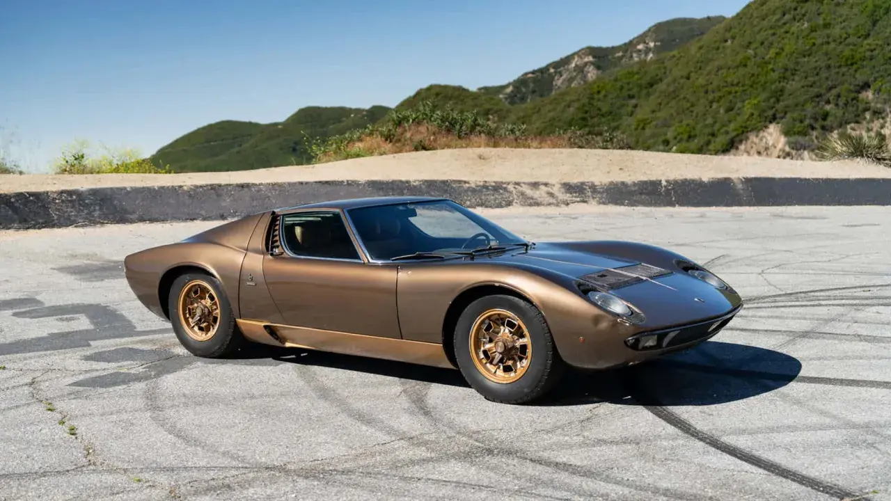 Ein brauner Lamborghini Miura S steht auf einer asphaltierten Straße mit Bergkulisse.
