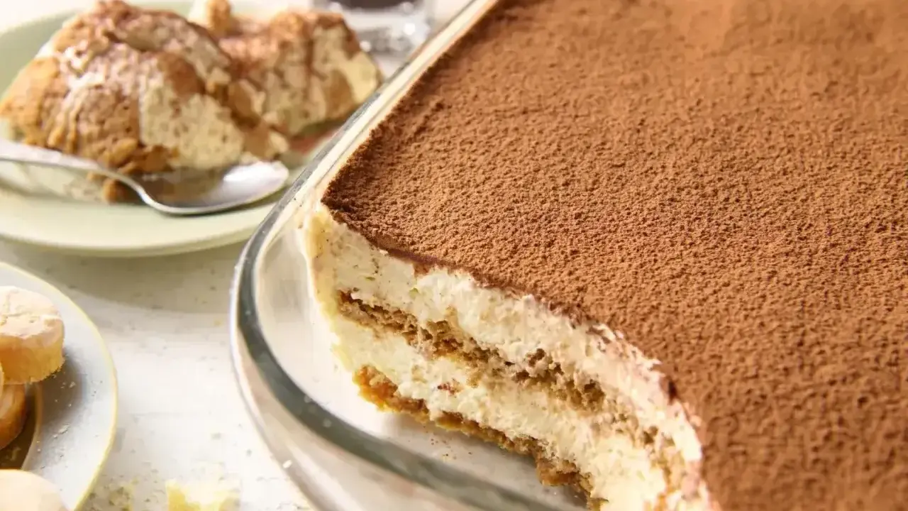Pyszne tiramisu bez jajek w szklanej formie, posypane kakao. Obok porcja deseru na talerzyku.