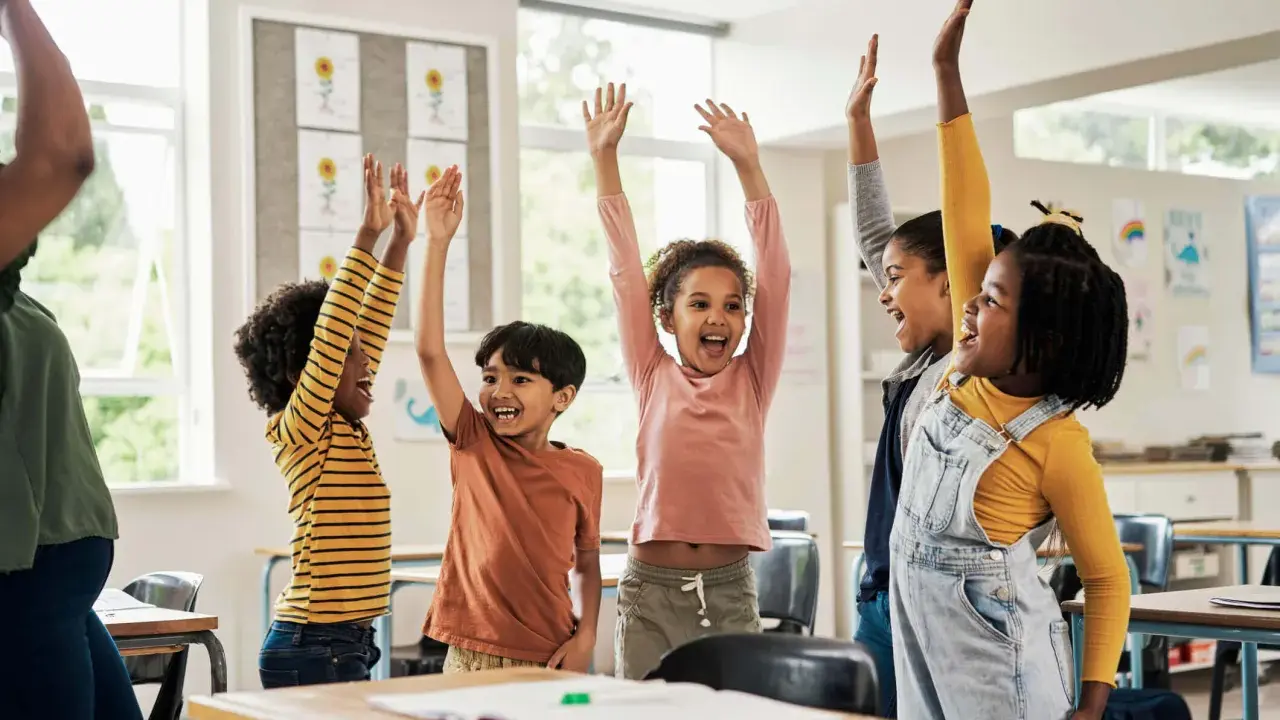 Fröhliche Kinder mit erhobenen Armen in einem Klassenzimmer. Sie nehmen begeistert an Kennenlernspielen für die Grundschule teil.