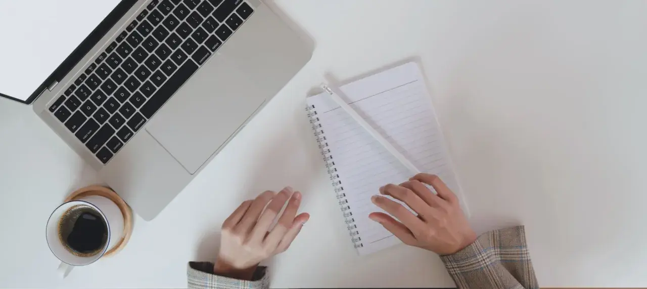 Ręce piszą notatki do CV po niemiecku przy laptopie, obok filiżanka kawy.