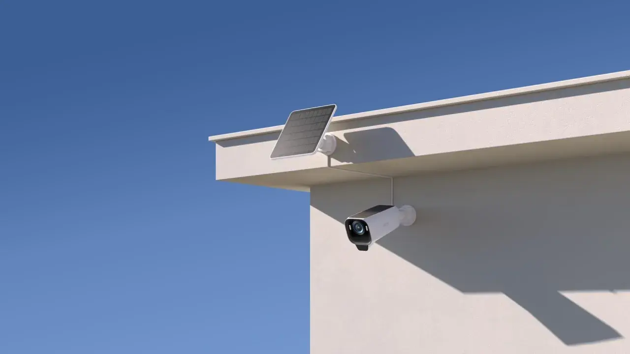 En eufycam 2 pro test visar en vit övervakningskamera med solpanel monterad på en vit vägg under en blå himmel.