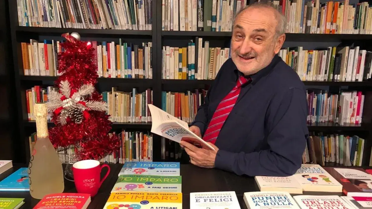 Daniele Novara, autore di libri per genitori, legge un suo libro tra scaffali pieni di volumi, con un albero di Natale e una bottiglia di spumante.