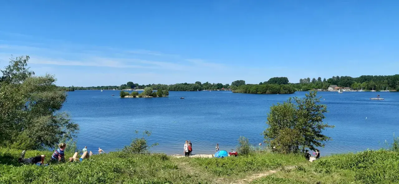 Sonniger Tag am Lippesee Paderborn. Menschen entspannen am Ufer, Segelboote gleiten über das Wasser, eine kleine Insel ziert die Mitte.
