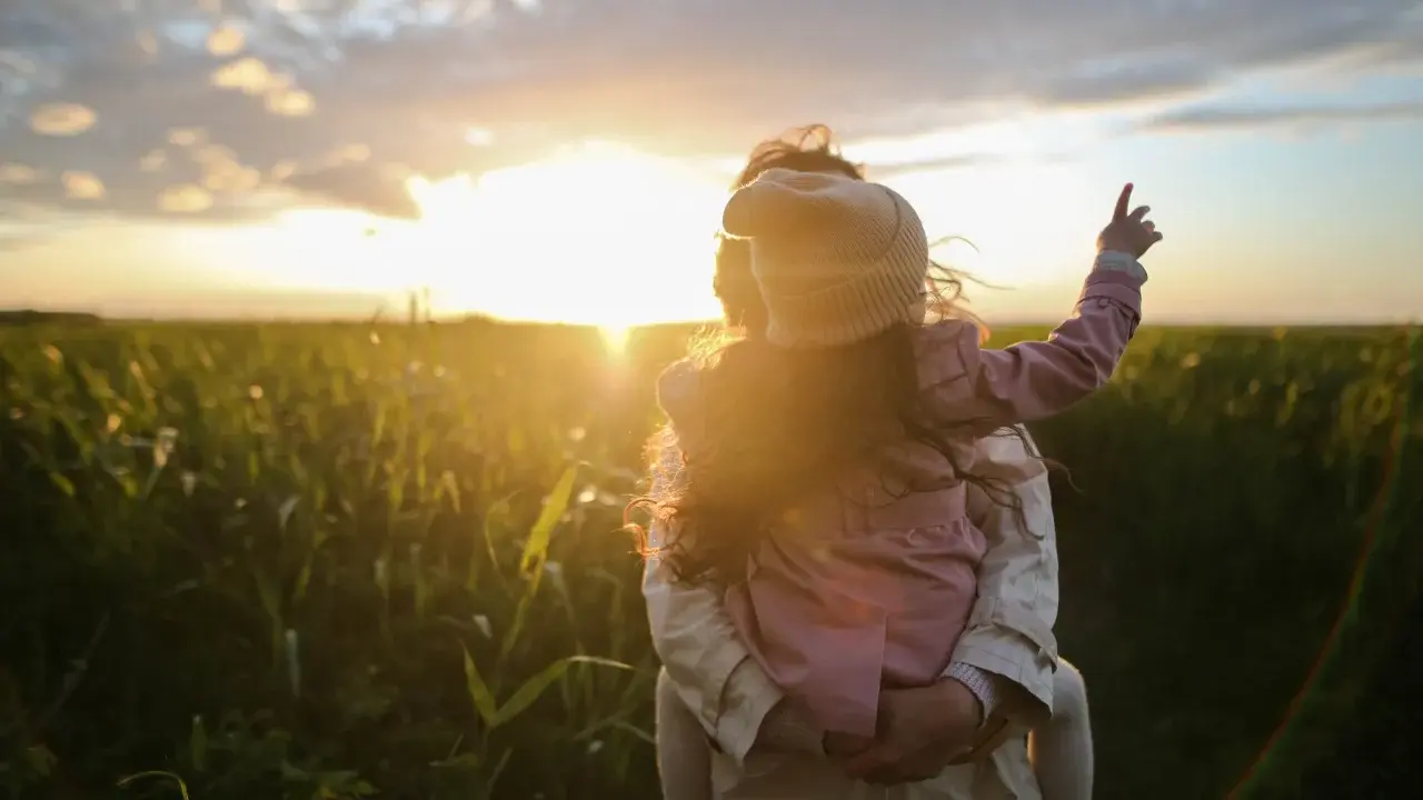 Bambina aggrappata alla schiena di un adulto, con il sole al tramonto che illumina un campo. Un'immagine di puro attaccamento affettivo.