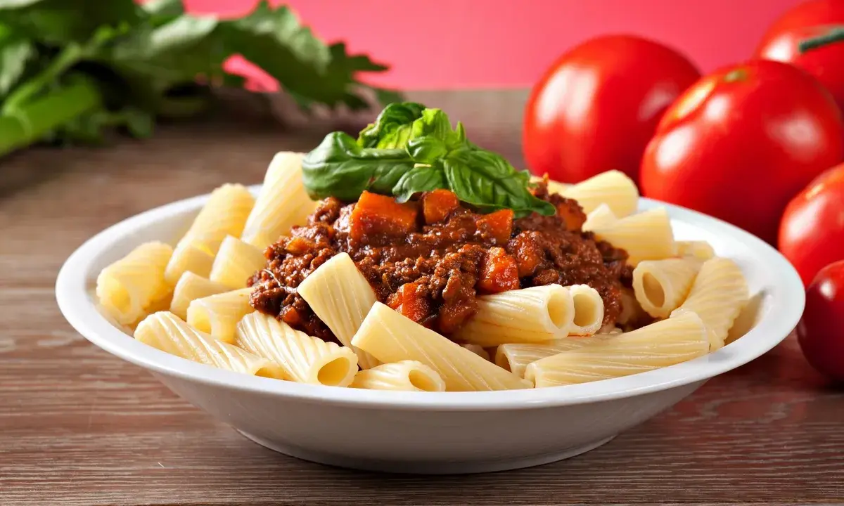 Pyszny makaron włoski rigatoni z sosem bolognese i świeżą bazylią, obok dojrzałe pomidory.
