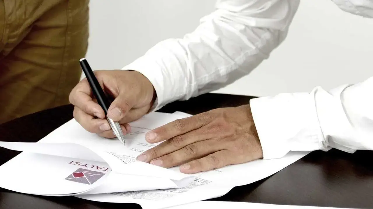 Mężczyzna podpisuje dokumenty, stając się ubezpieczającym dla ubezpieczonego. Na papierze widoczne logo firmy.