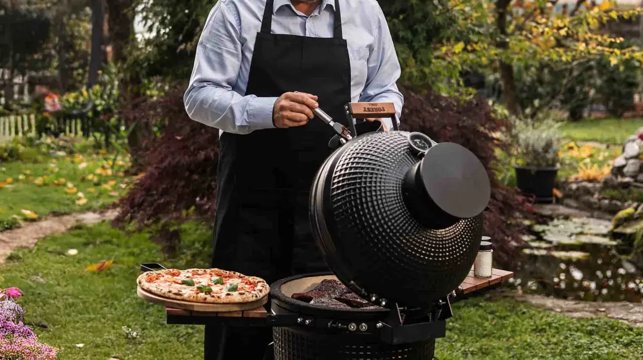 Un homme en tablier noir prépare une pizza et des steaks sur un kamado, un barbecue céramique polyvalent, dans un jardin verdoyant.