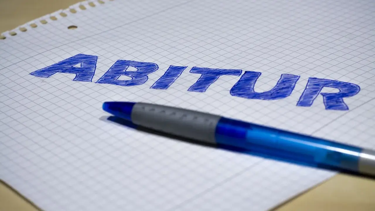 Blauer Stift liegt auf kariertem Papier, auf dem das Wort "Abitur" geschrieben ist. Der **Abitur**-Abschluss rückt näher.