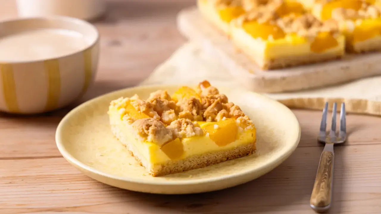 Ein Stück saftiger Pfirsich Blechkuchen mit Vanillepudding und Streuseln auf einem gelben Teller, daneben eine Gabel.