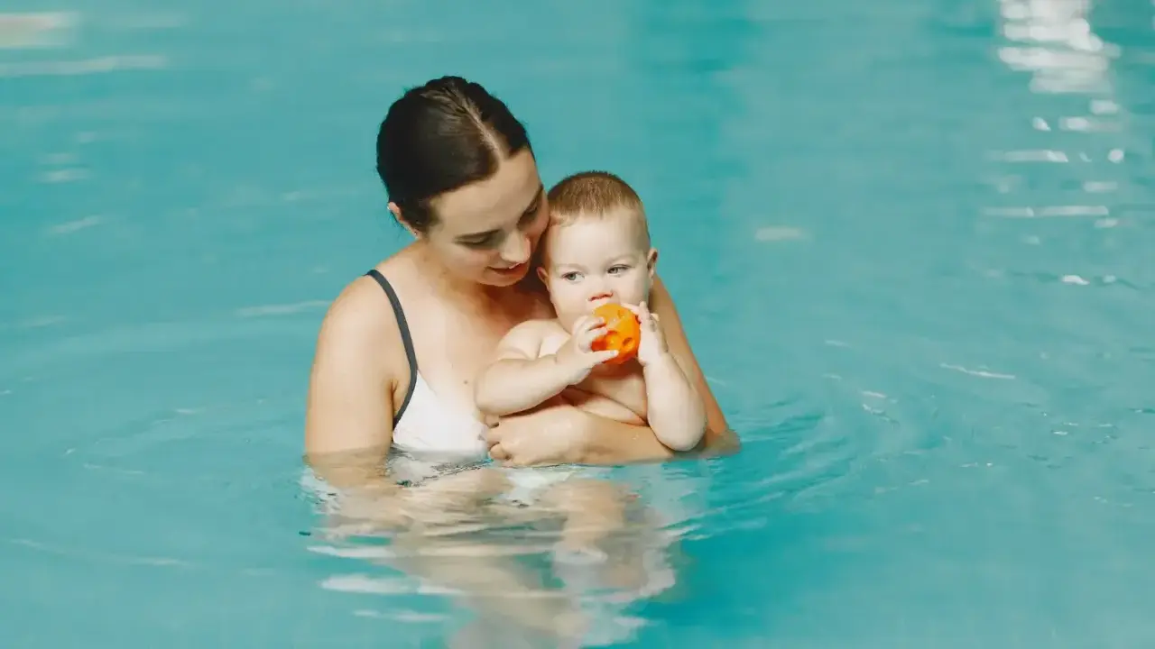 Mama trzyma roczne dziecko na basenie. Maluch bawi się pomarańczową piłeczką.