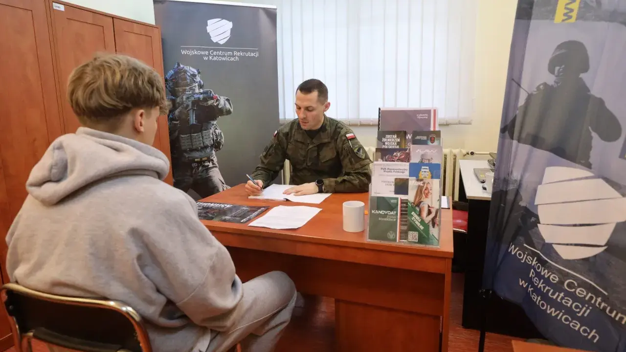 Młody mężczyzna rozmawia z żołnierzem w punkcie rekrutacyjnym. Dyskutują o możliwościach związanych z obowiązkową służbą wojskową.