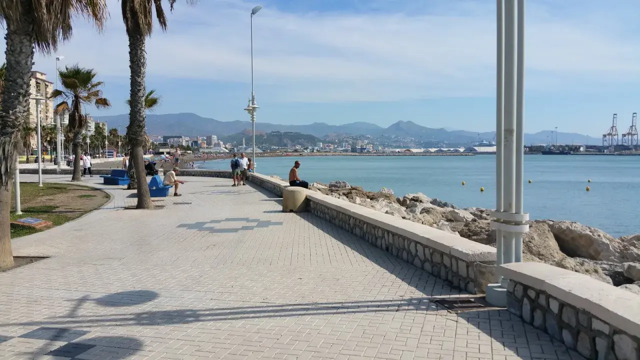Ein Mann sitzt am Ufer der playa de la misericordia und blickt auf das Meer. Palmen säumen die Promenade, im Hintergrund die Stadt und Berge.