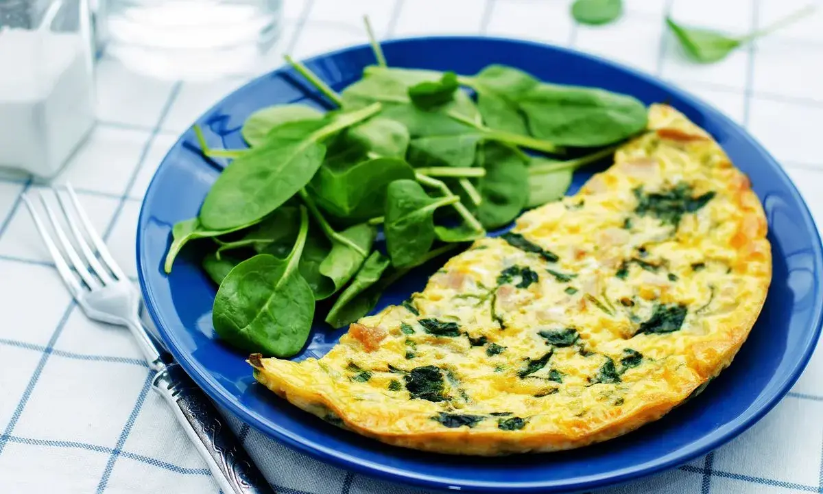Frittata di spinaci dorata su piatto blu, accompagnata da spinacini freschi. Un pasto leggero e gustoso.