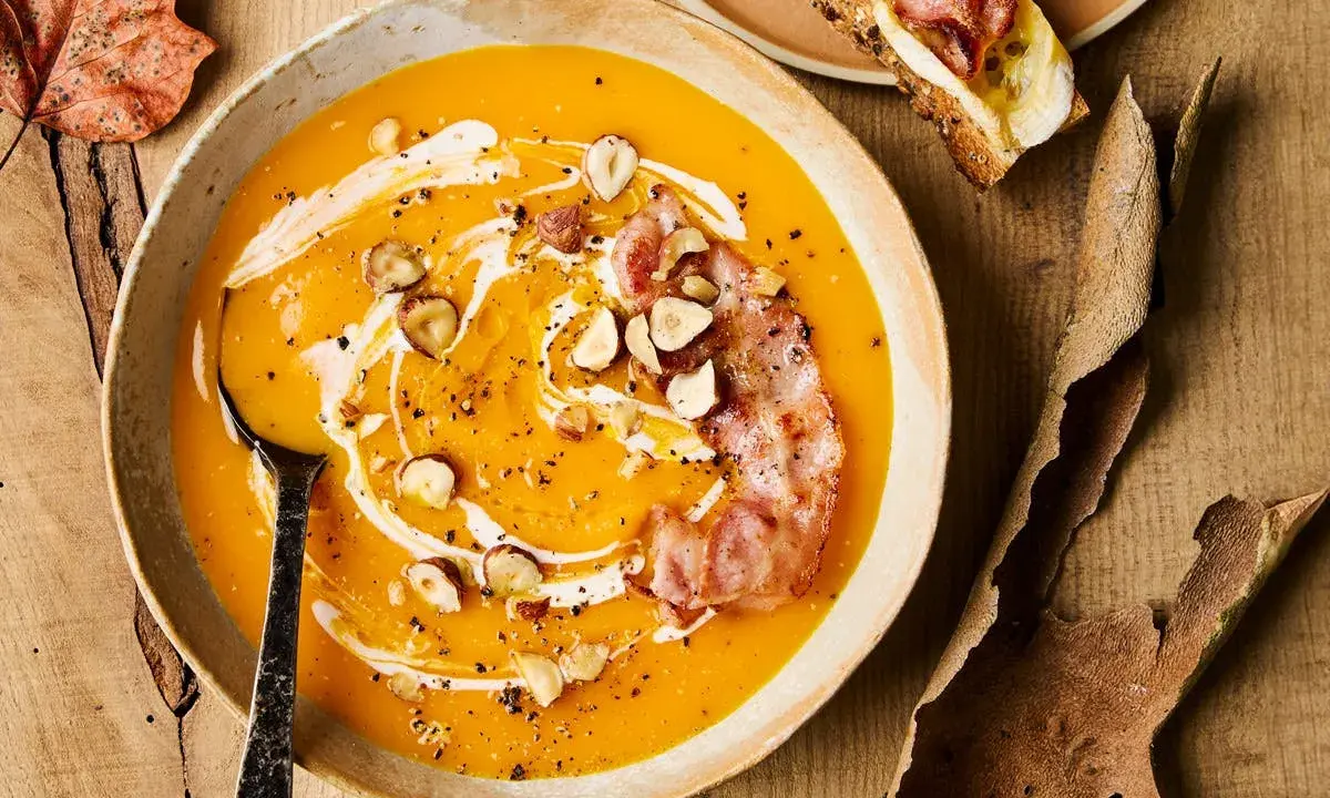 Velouté de courge onctueux, garni de noisettes croquantes, d'une touche de crème et d'une tranche de bacon croustillant. Un délice automnal qui évoque le beurre de courge.