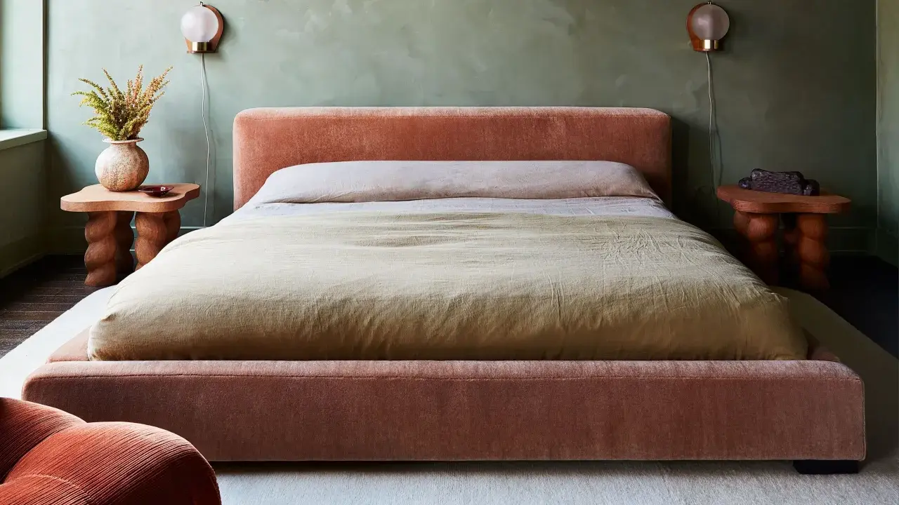 Cama king size tapizada en terciopelo rosa, con mesitas de noche de madera y lámparas de pared.