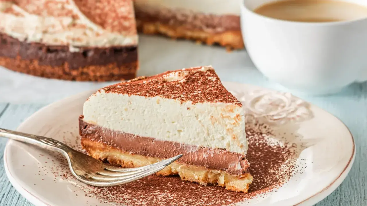 Pyszna tarta z mascarpone i czekoladą, posypana kakao, czeka na widelczyk. Obok filiżanka kawy.