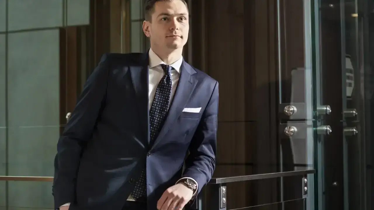 Mężczyzna w granatowym garniturze i krawacie stoi oparty o balustradę.